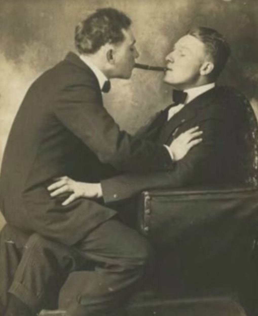 Two men in a B&W photograph. One is sitting on the other's lap and they both are pressing their cigars together from their mouths. Looks to be late 1800s to early 1900s.